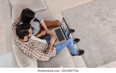 Young Indian Couple Using Laptop Having Video Call Family Virtual Meeting At Home. Smiling Ethnic Husband And Wife Sitting On Couch Talking With Distance Grandparent In Online Video Chat. Top View