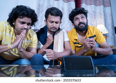 Young Indian cricket sport fans praying for supporting team to win while watching live match on mobile phone at home - concept of tense, curious emotional or mental stressful game