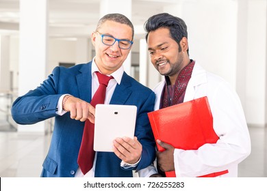 Young Indian doctor with businessman talking in hospital hallway - Salesman holding pad showing health care equipment at asian medical director - Concept of sales, business, deals with multiracial men