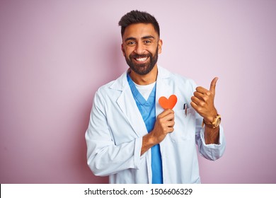 Young Indian Doctor Man Holding Paper Heart Standing Over Isolated Pink Background Happy With Big Smile Doing Ok Sign, Thumb Up With Fingers, Excellent Sign