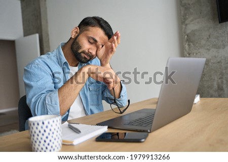 Young indian eastern tired exhausted business man rubbing eyes sitting in modern home office with laptop on desk. Overworked burnout academic Hispanic student with glasses in hand feeling eyestrain.