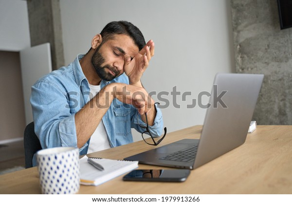 Young indian eastern tired exhausted business\
man rubbing eyes sitting in modern home office with laptop on desk.\
Overworked burnout academic Hispanic student with glasses in hand\
feeling eyestrain.