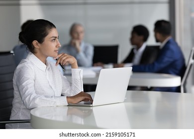 Young Indian employee thinking over business task looks thoughtful sit at desk with laptop, diverse colleagues on background negotiating. Professional occupation worker workflow in workspace concept - Shutterstock ID 2159105171