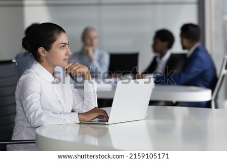 Young Indian employee thinking over business task looks thoughtful sit at desk with laptop, diverse colleagues on background negotiating. Professional occupation worker workflow in workspace concept