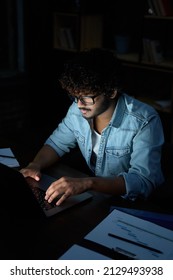 Young Indian Ethnic Business Man Developer Or Web Designer Wearing Glasses Typing On Laptop Distance Working Online Late At Night At Home Or In Dark Office Using Laptop Sitting At Workplace Desk.