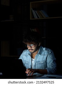 Young Indian Ethnic Businessman Developer Or Web Designer Wearing Glasses Typing On Laptop Distance Working Online Late At Night At Home Or In Dark Office Using Laptop Sitting At Workplace Desk.