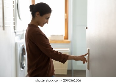 Young Indian ethnicity female push button turns off on light inside modern domestic laundry room, adjusting thermostat for controlling air conditioner and heater inside house. Climate control concept - Shutterstock ID 2070561506