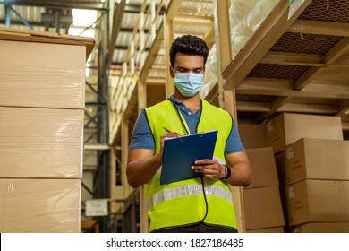 Young Indian Factory Warehouse Worker Wearing A Protective Face Mask While Working In Logistic Industry Indoor. 30s Man Checking Item Stock Order During Coronavirus Covid 19 Pandemic Outbreak