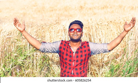 Young indian farmer raising arms in wheat field praising god for the harvest - African american man hands up at sky inside golden corn field on sunny summer day - Concept of successful hard farm work