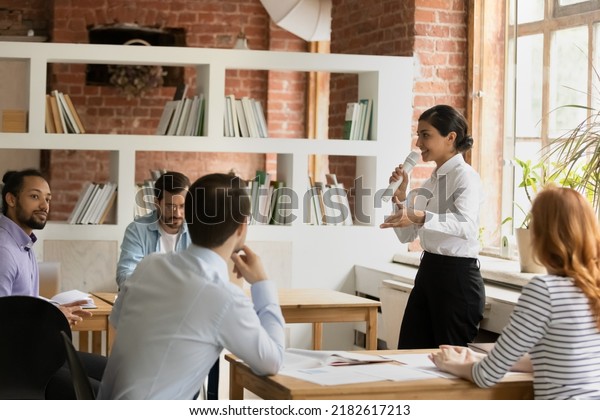 Young Indian female business coach makes speech\
into microphone for group of diverse company staff employees during\
educational seminar conference in modern office. Corporate training\
event concept