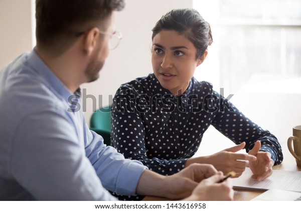 Young indian female mentor coach worker talking to\
male coworker teaching intern having business conversation with\
workmate, serious hindu manager helping colleague discussing new\
project in office
