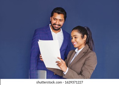 Young Indian Female Professional Explaining Contract Details To Tall Indian Handsome Man In Business Suit, Isolated Over Blue Studio Background