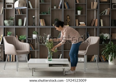 Young Indian female renter or tenant decorate cozy modern home or apartment. Millennial mixed race woman take care of flowers, involved in living room decoration. Interior design, rent concept.