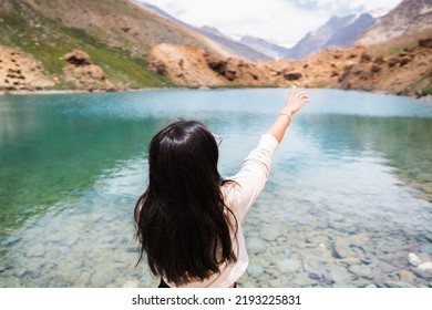Young Indian female traveler pointing her finger towards Himalayan mountains while standing at clear blue water lake Deepak Tal, Himachal Pradesh, India. Travel and holiday concept. Summer in mountain