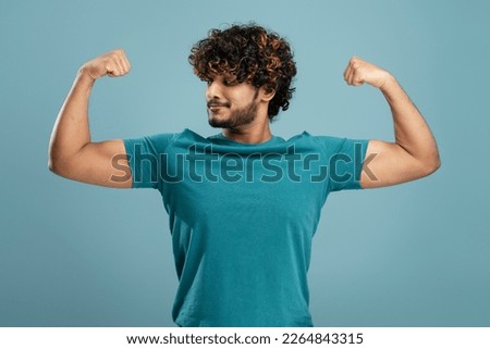 Young Indian fitness man wearing casual blue t-shirt over isolated background showing arms muscles and smiling proud. Fitness concept. Super Power. Young strong bearded Asian sportsman showing biceps.