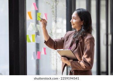 young indian girl asian woman entrepreneur of small company putting a adhesive sticky notes in glass wall in office during analyzing strategy team meeting formulating business strategies. - Powered by Shutterstock