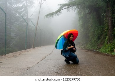 Young indian girl with colorful umbrella sitting on foggy street with trees in the background. Shows a perfect holiday vacation image of trekking in the himalayas in himachal pradesh