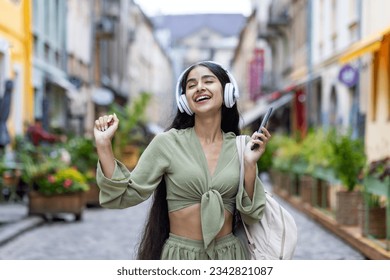 A young Indian girl is dressed in headphones, holding a phone in her hand. Closing his eyes dances on the streets of the city, listening to music, enjoying. - Powered by Shutterstock