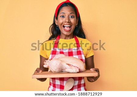 Young indian girl wearing cook apron and holding chicken smiling and laughing hard out loud because funny crazy joke. 