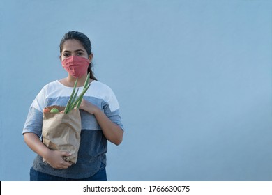 Young Indian girl wearing hand stitched cotton mask holding grocery bag with vegetables during Covid 19 outbreak. Budget buying the necessities. Grey background with copy space - Shutterstock ID 1766630075