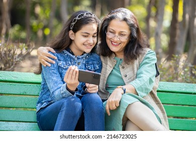 Young Indian Granddaughter Holding Smart Phone Showing Something To Her Grandmother While Sitting On Bench At Park. Happy Mature Old Asian Woman With Girl Child Using Mobile Phone Together Outdoor.