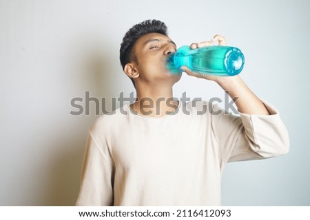 An young Indian guy drinking from a bottle of water against a white background
