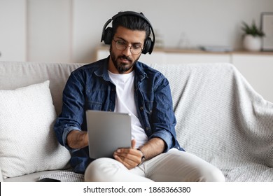 Young Indian Guy Spending Time At Home With Digital Tablet And Wireless Headphones, Relaxing On Comfortable Couch In Living Room, Millenial Western Man Checking New Application Or Browsing Internet