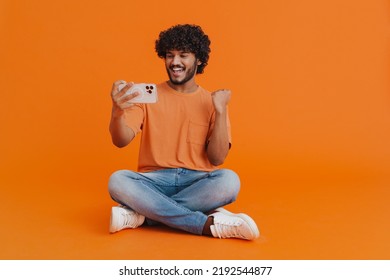 Young indian handsome curly enthusiastic man holding his phone celebrating victory in mobile game with raised fist while sitting in lotus pose over isolated orange background - Shutterstock ID 2192544877
