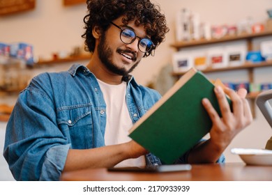 Young indian handsome man in glasses reading book at cafe