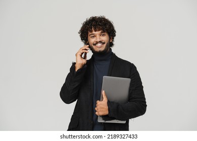 Young Indian Handsome Man Talking To The Phone With Laptop Looking On Camera On Grey Isolated Background