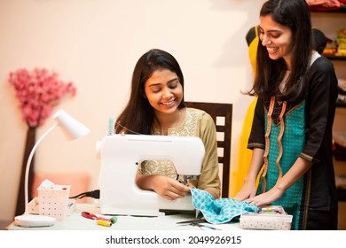 Young indian happy women,tailor,fashion designer,using sewing machine,talking to each other and smiling,sit behind her desk in her garment tailoring workshop,atelier or studio.
