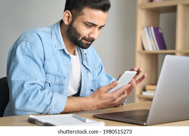 Young Indian Hispanic Businessman Using Smartphone And Computer Working Remotely From Modern Home Office. Latin Student Having Virtual Training On Cellphone Texting Using Online Tech Applications.