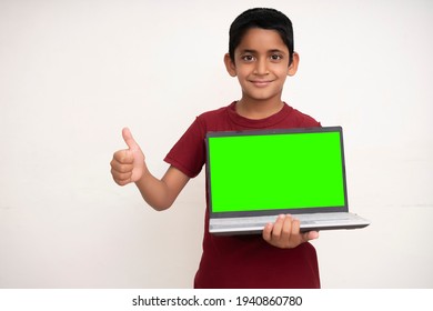 Young Indian Kid Holding A Laptop With Green Screen In His Hands And Showing Thumbs Up To The Camera. Standing On A White Isolated Wall With Copy Space.