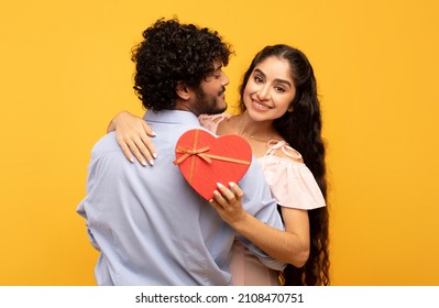 Young Indian Lovers Hugging On Yellow Background, Lady Holding Heart Shaped Gift Box And Smiling At Camera. Loving Couple Celebrating Valentine's Day Or Anniversary Together