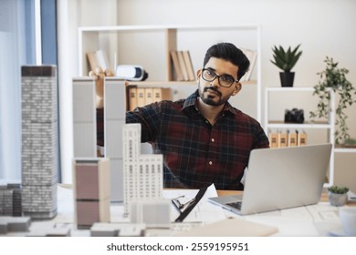 Young Indian male architect working on city planning with laptop and scale models. Focused professional at work in modern office setting. Concept of architecture, creativity, urban planning. - Powered by Shutterstock