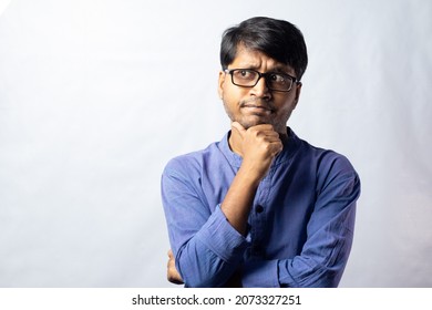 A Young Indian Male In Blue Ethnic Dress And Glasses Posing With Hand Under Chin On White Background