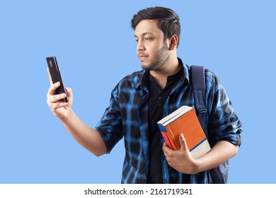 Young Indian Male Collage Student Using Mobile Phone With Books Standing Against Single Color Blue Background