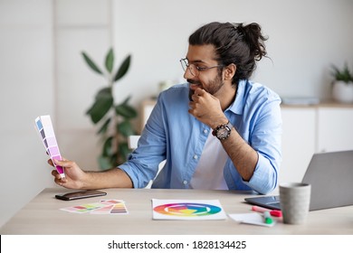 Young indian male designer holding and looking at colour swatches, working at desk in creative office, choosing interior design, stylish western man checking color gamma for new project, free space