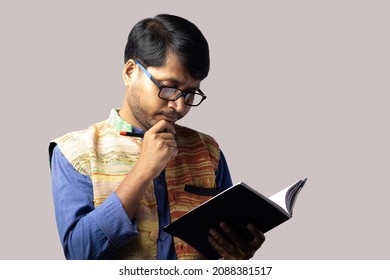 A Young Indian Male In Ethnic Dress Thinking With Pen And Notebook On Gray Background