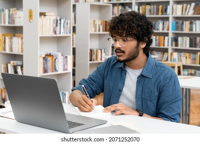 Young Indian Male Student Watching Education Webinar Using Laptop Writing Notes Doing Research On Internet Sitting At High School Library. Digital Technology, Communication And Studying