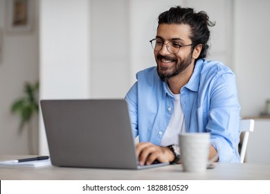 Young Indian Male Writer Using Laptop Computer, Sitting At Desk In Home Office, Handsome Western Man In Eyeglasses Typing On Keyboard And Looking At Screen, Enjoying Working Remotely, Selective Focus - Powered by Shutterstock