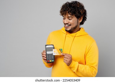 Young Indian man 20s he wear casual yellow hoody hold wireless modern bank payment terminal to process acquire credit card isolated on plain grey background studio portrait. People lifestyle portrait - Shutterstock ID 2254053409