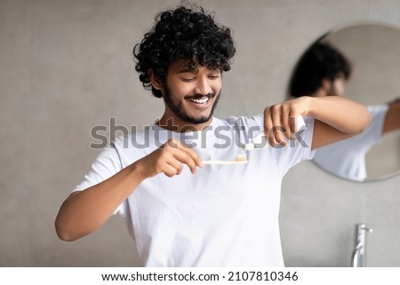 Young indian man applying toothpaste on toothbrush, brushing teeth in the morning, standing in bathroom, copy space. Toothcare and daily oral hygiene routine concept
