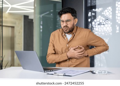 A young Indian man in a brown shirt is sitting at the desk in the office and is bent over from pain in the heart, holding his hands to his chest, feeling severe pain. - Powered by Shutterstock