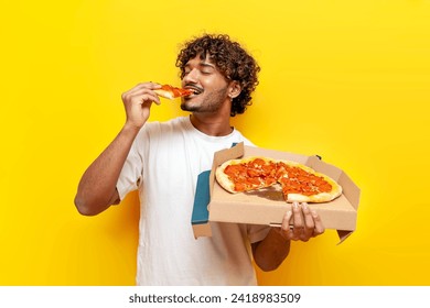 young indian man eating delicious pizza on yellow isolated background, curly guy student holding pizza box and biting a piece of fast food