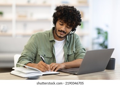 Young indian man having online training or attending webinar, sitting at workdesk, using laptop and headset, taking notes and smiling, home interior, copy space. Online education concept - Shutterstock ID 2190346705