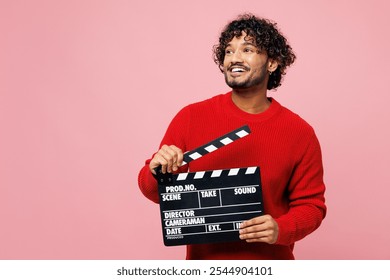 Young Indian man he wears red sweater casual clothes hold in hand classic black film making clapperboard look aside isolated on plain pastel light pink background studio portrait. Lifestyle concept