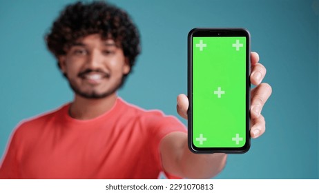 Young indian man holding a phone with green screen, showing a blank chromakey screen in coral t-shirt on blue studio background. - Powered by Shutterstock