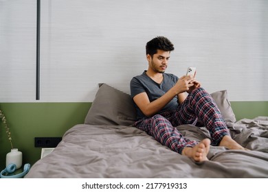 Young Indian Man Looking On His Phone Sitting On The Bed At Home