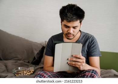 Young Indian Man Scrolling Tablet Sitting On Bed With Bowl Of Nuts At Home